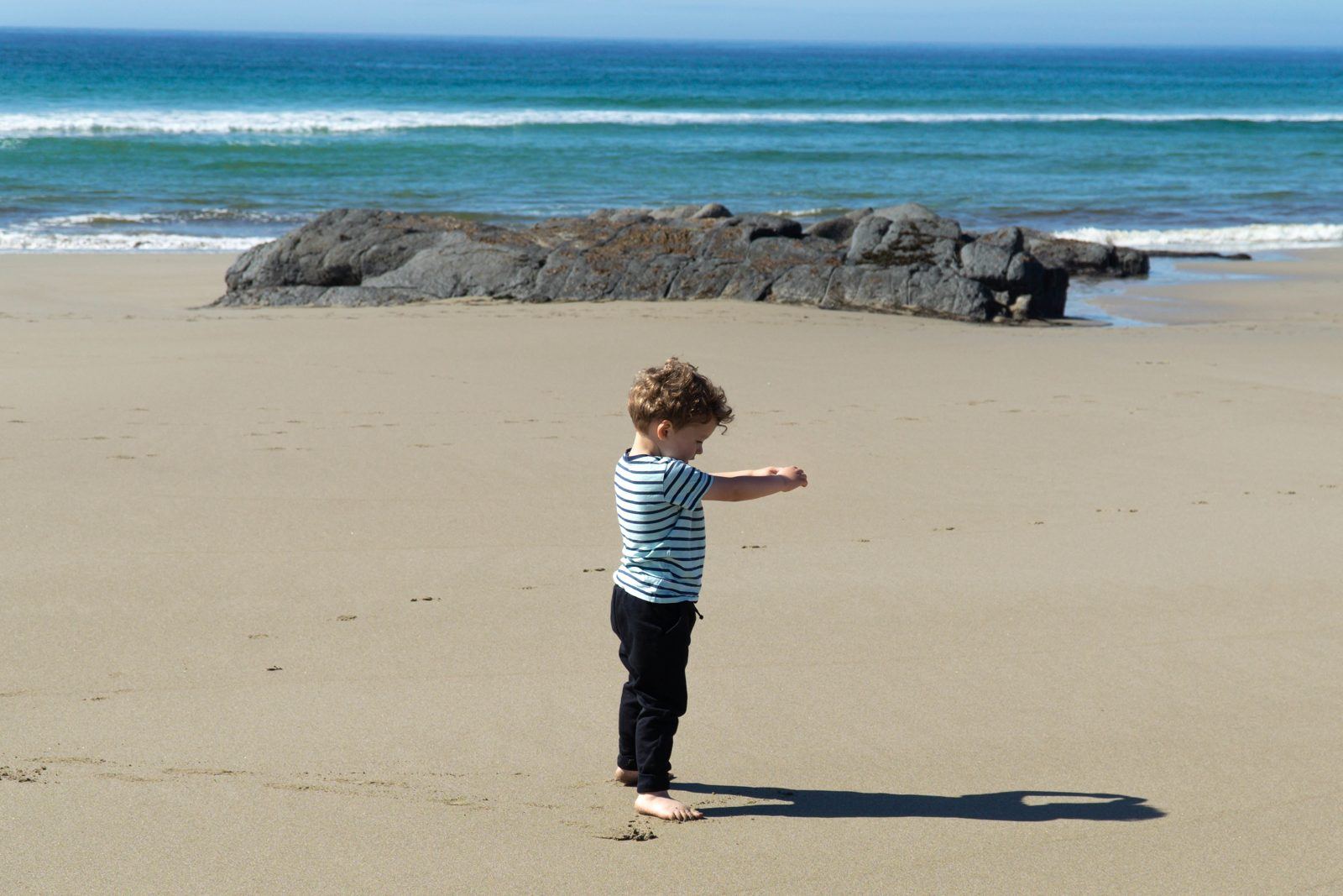 A young child in a striped top playing on a wide sandy beach, with rocks and surf behind